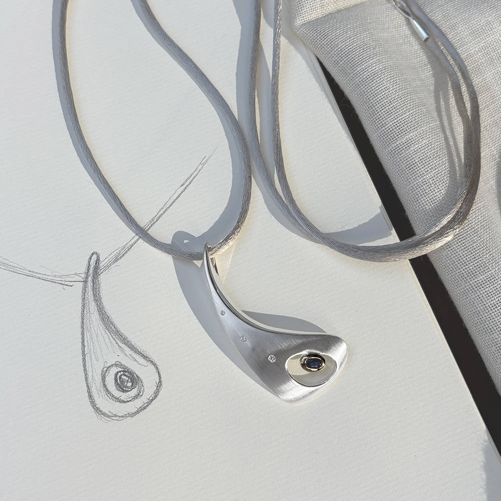 Outback Fossil Pendant in silver with sapphire set in 18ct yellow gold and diamonds, resting on a design sketch. Sculptural pendant inspired by the Australian outback, highlighting its organic form and refined craftsmanship.